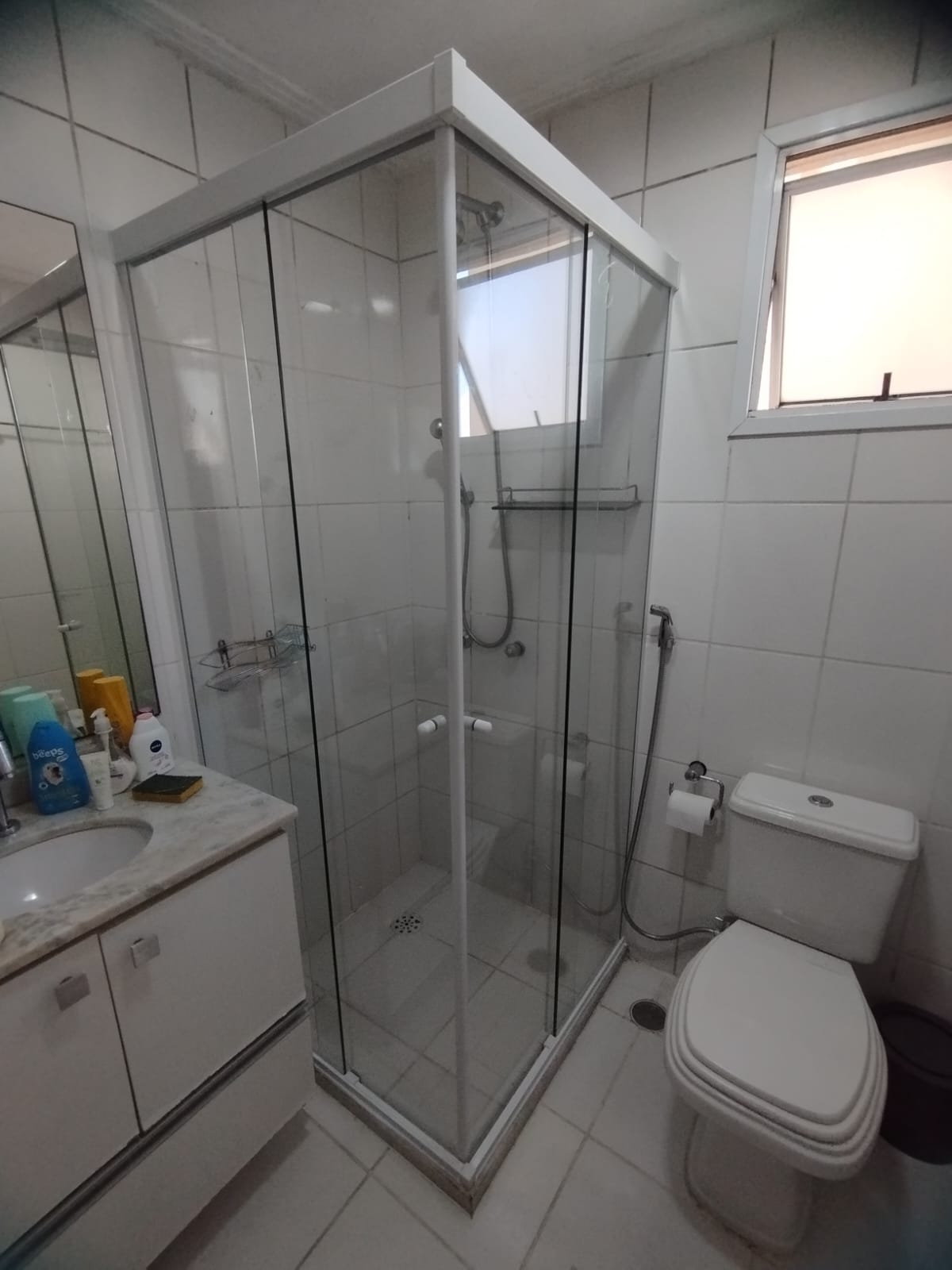 um banheiro com banheira de vidro, pia com espelho e toilette, todas com revestimento de azulejos brancos — t